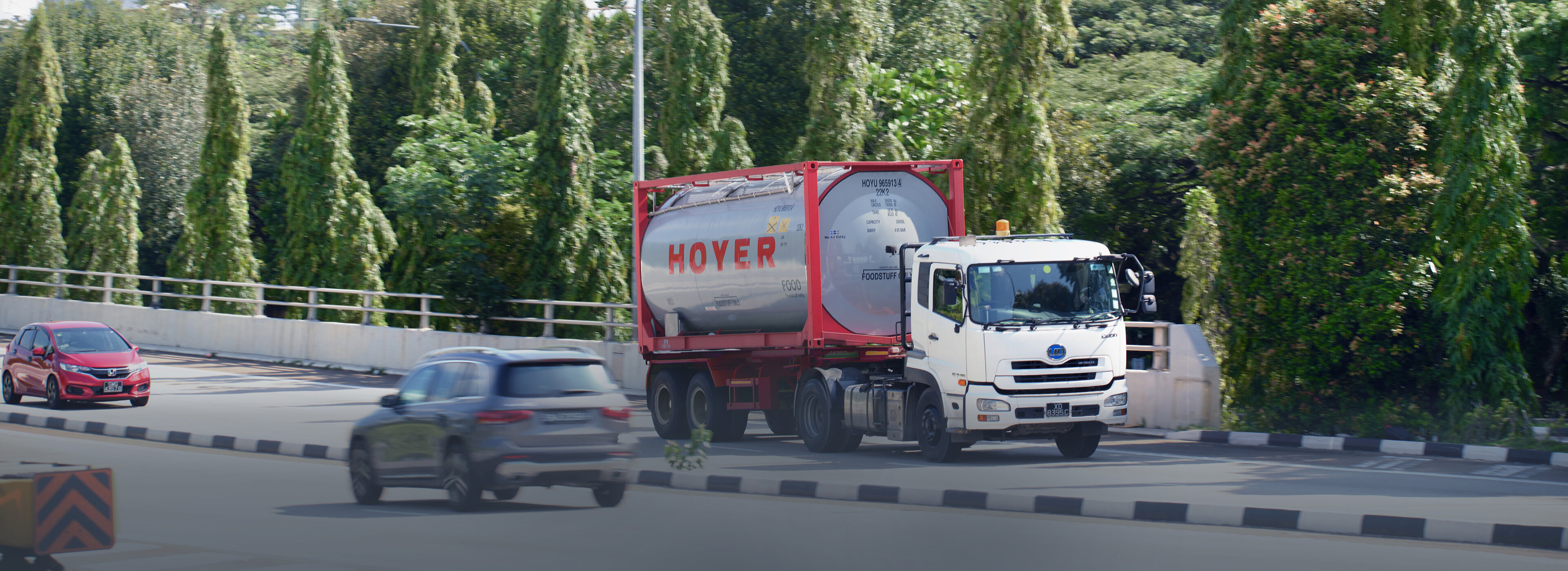 A white truck with a HOYER food tank container drives on a tree-lined road, two cars in the background