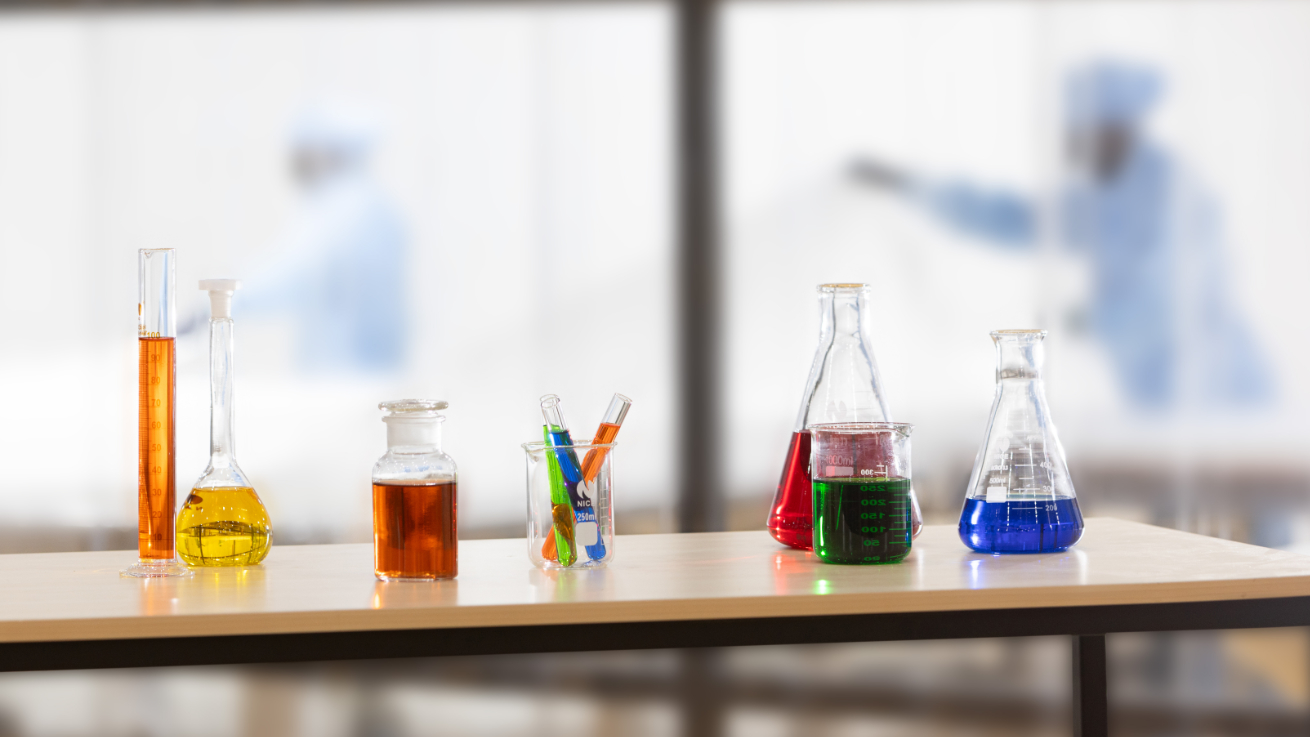 Laboratory environment with glass containers filled with colorful liquids, in the background two people in protective clothing in flexitank production