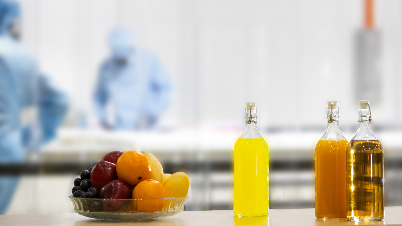 Fruit bowl with apples, oranges and grapes next to glass bottles with liquids, in the background employees in protective clothing in flexitank production