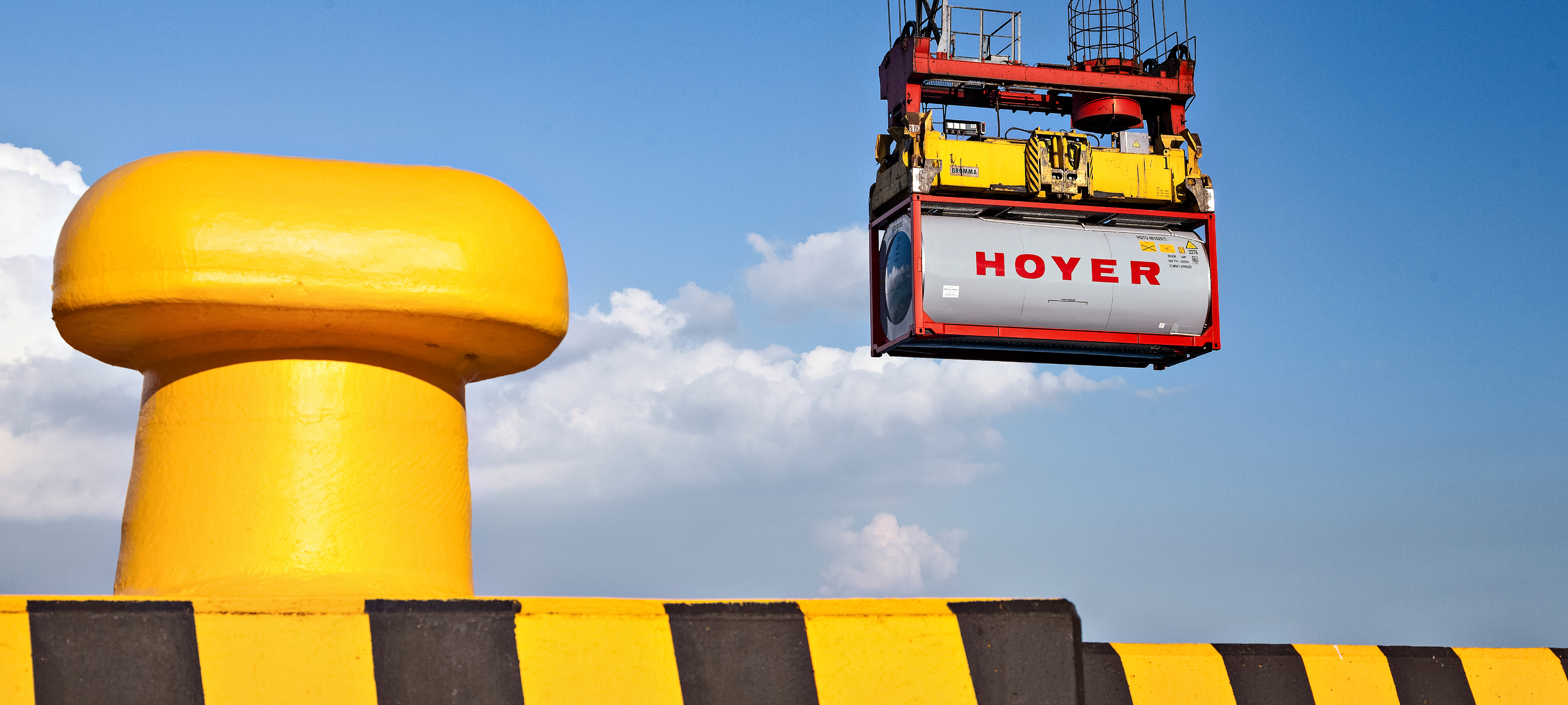 HOYER chemical tank container being lifted by a crane, with safety barriers and a clear sky in the background