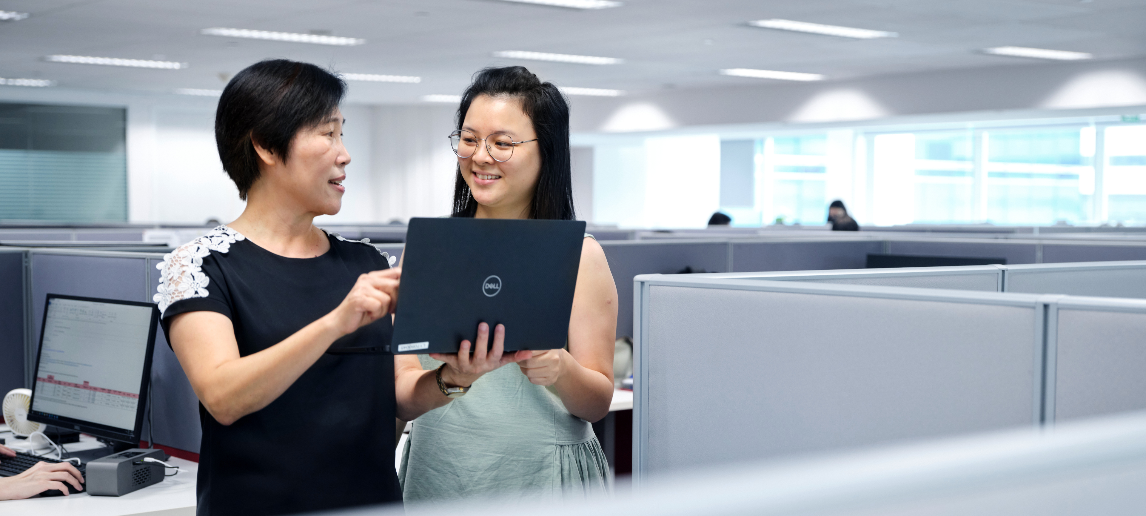 Two office colleagues reviewing content on a laptop in a modern workspace with cubicles and computer workstations