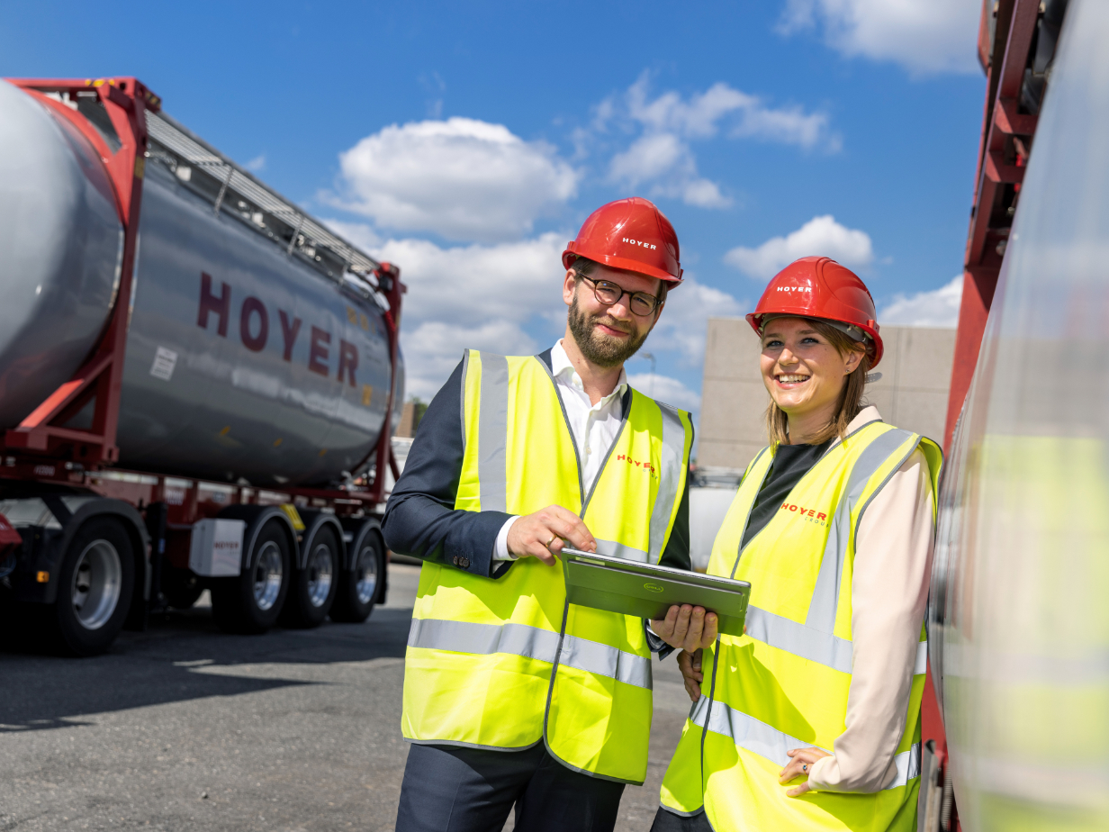 Two employees wearing high-visibility vests and red helmets standing next to a HOYER chemical tank container and look at a tablet together