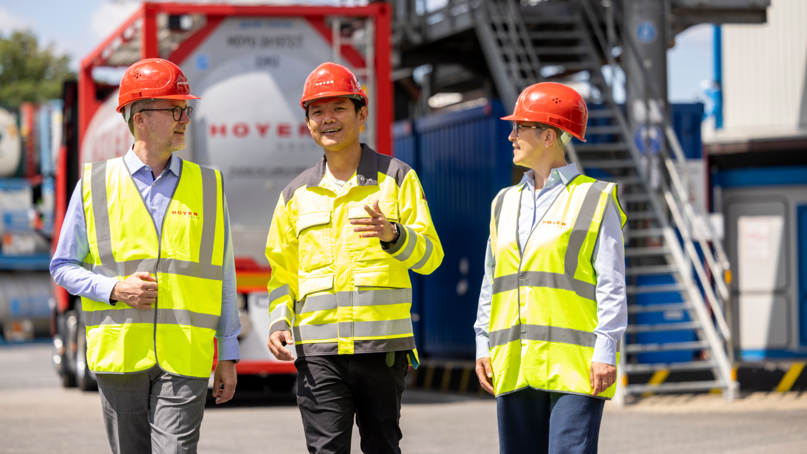 Three employees wearing high-visibility vests and red helmets standinging in front of a HOYER chemical tank container with industrial structures in the background