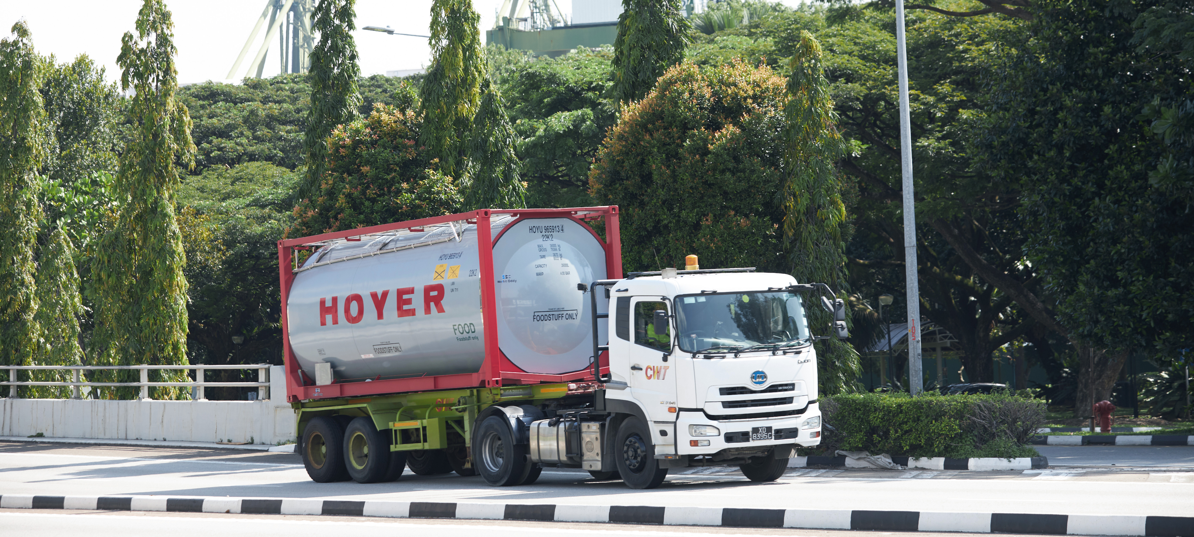 HOYER food tank container on white truck with red frame, driving past trees and industrial structures under clear sky