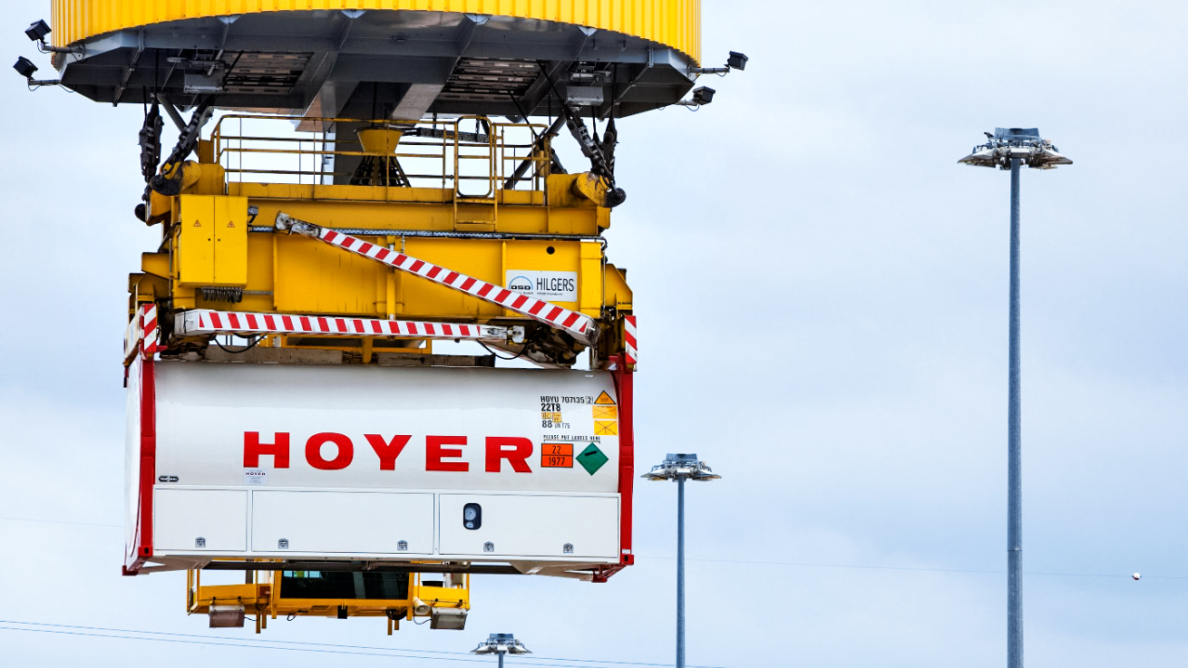 HOYER gas container with hazardous goods labels being hoisted by yellow crane with red-white safety stripes at logistics terminal