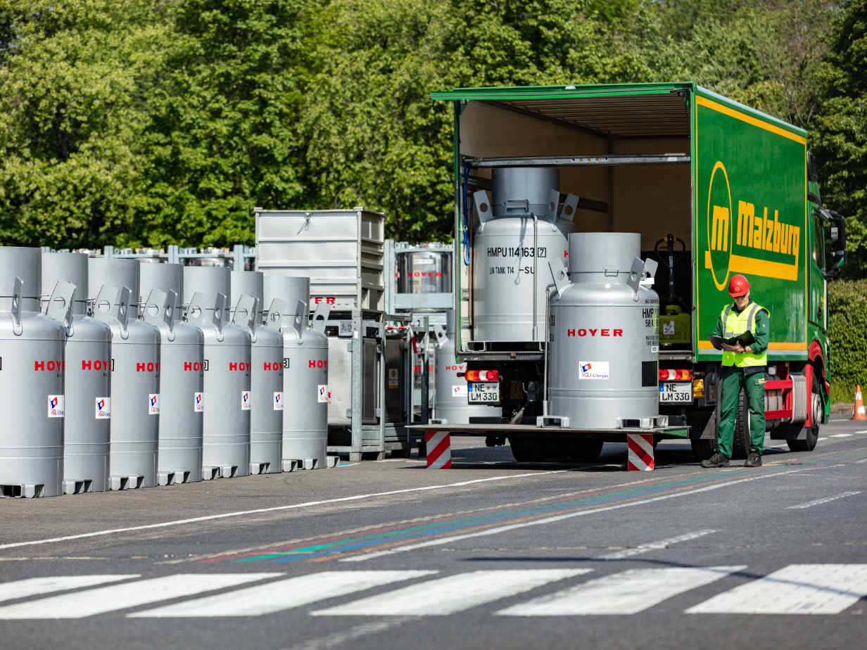 Worker in red helmet and green vest checking loading of HOYER-labeled IBCs into green truck at outdoor site