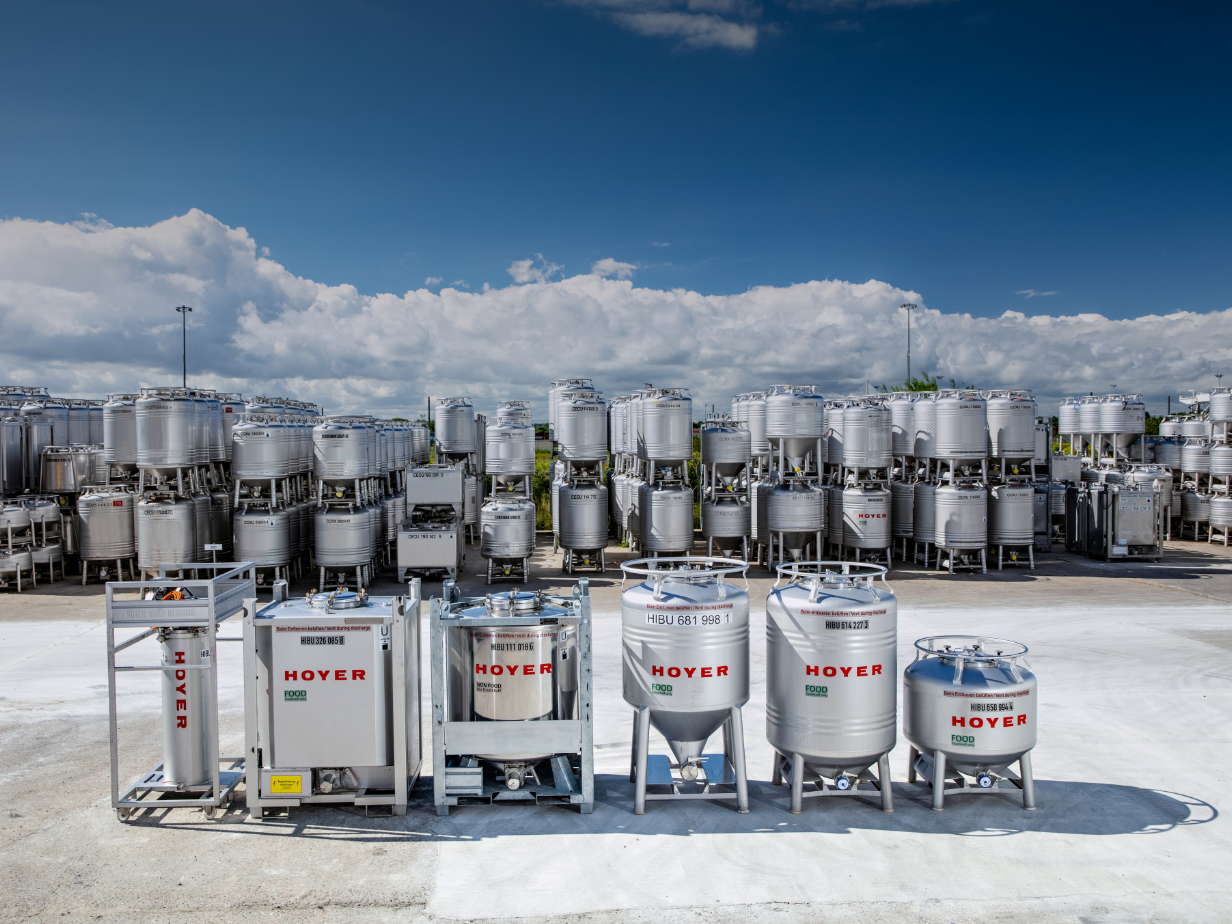 Outdoor storage yard filled with HOYER-labeled IBCs in various sizes, stacked on metal racks under a partly cloudy sky