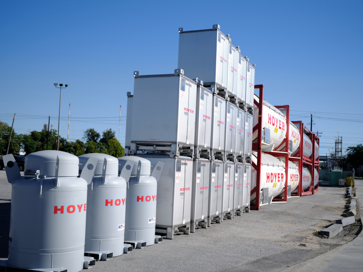 Stacked IBCs and HOYER tank containers in outdoor depot area with trees, power lines, and blue sky in the background