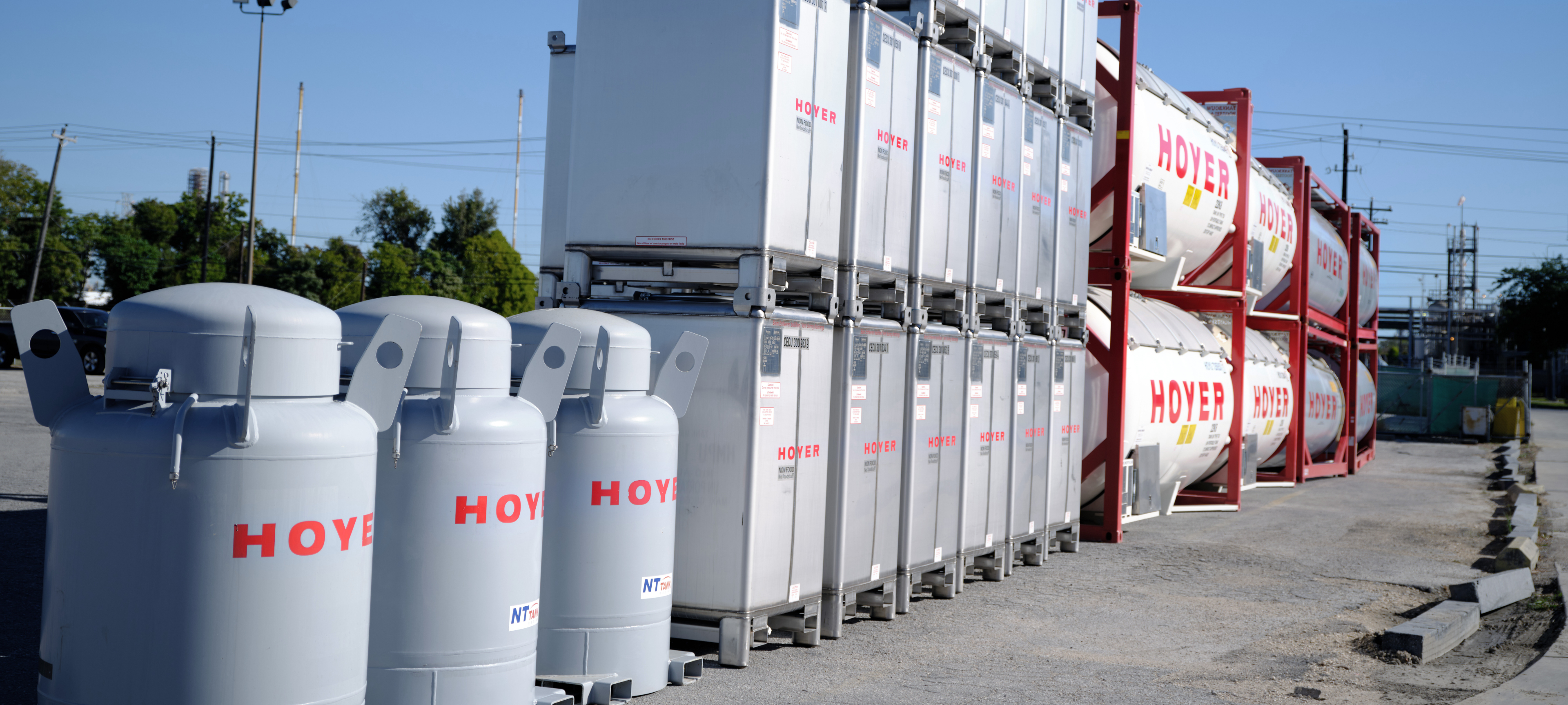 Stacked IBCs and HOYER tank containers in outdoor depot area with trees, power lines, and blue sky in the background