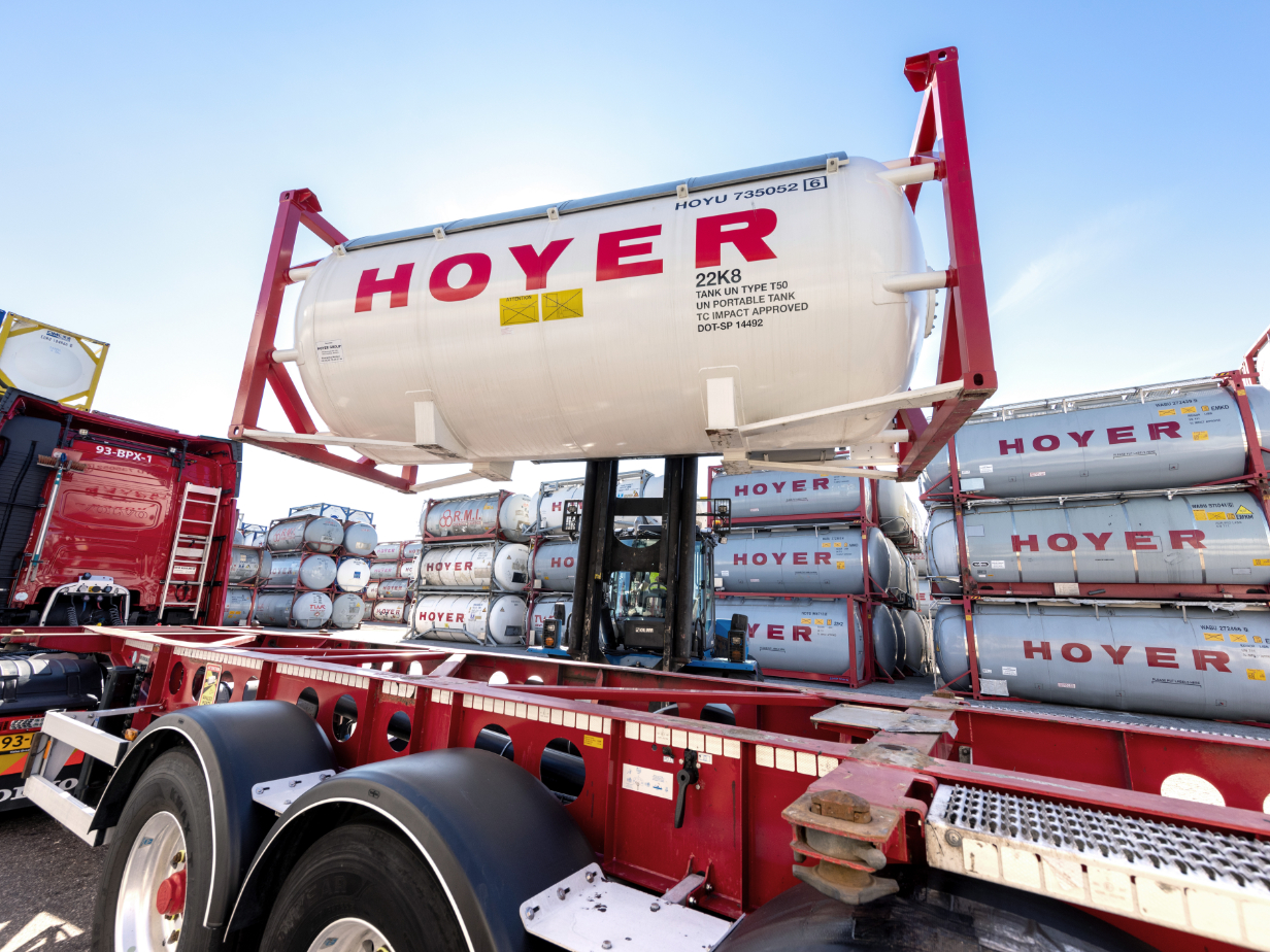 Forklift loading HOYER gas tank container onto truck chassis at depot site with stacked containers in the background
