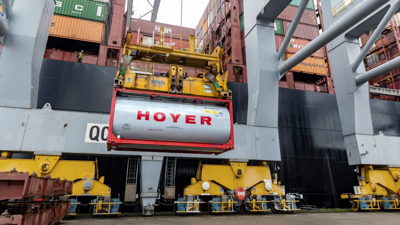 HOYER tank container being lifted by crane onto cargo ship for overseas transport, with stacked containers in the background