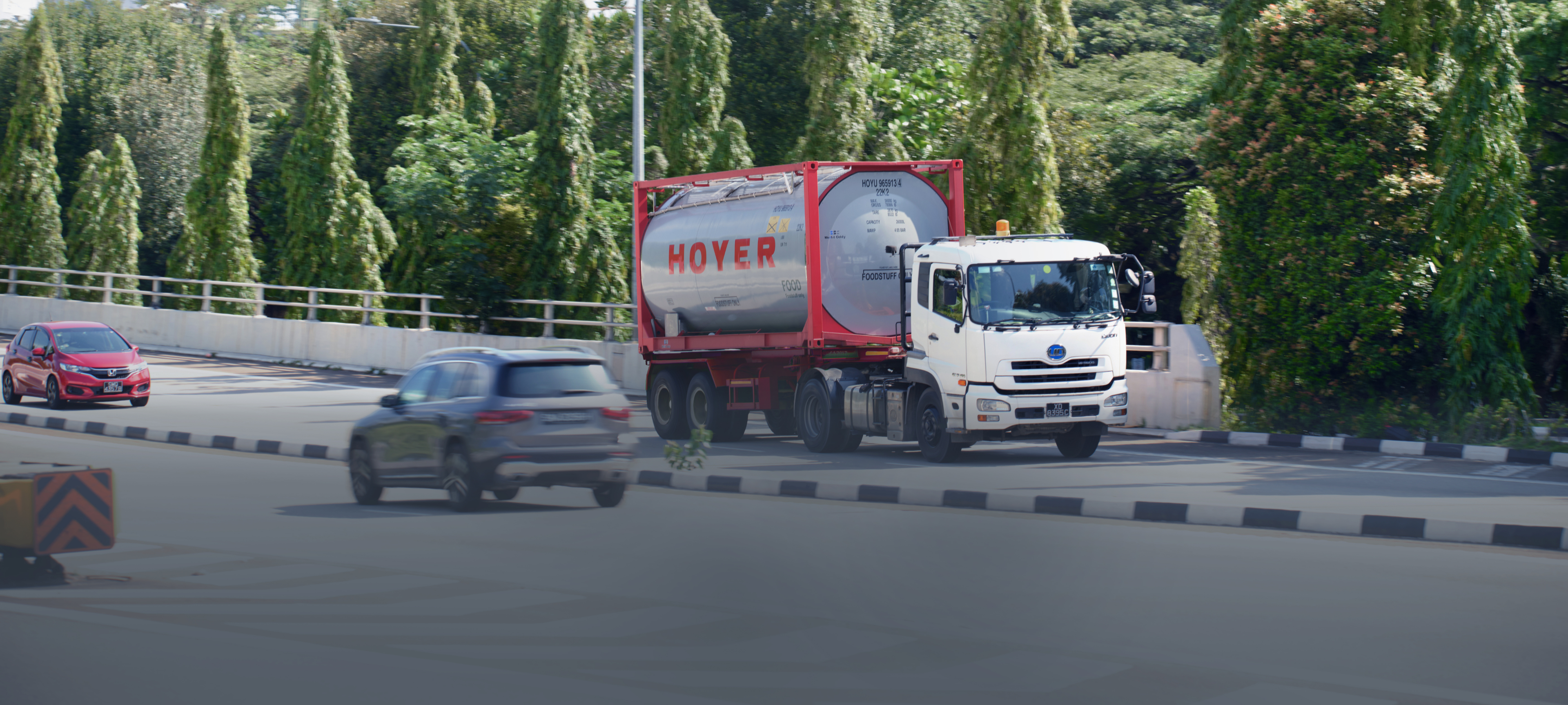 一辆装载HOYER食品罐式集装箱的白色卡车行驶在林荫大道上，背景中有两辆汽车