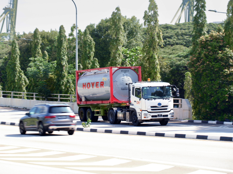 A white truck with a HOYER food tank container drives on a road surrounded by trees, two cars in the background